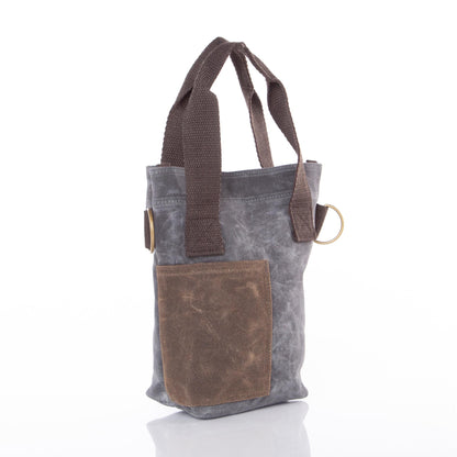 Gray waxed canvas wine bottle tote with brown leather pocket, brown fabric handles, and brass ring detail; no visible monogram or embroidery