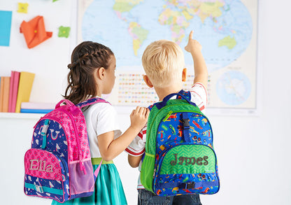 Two children wearing personalized Stephen Joseph Rainbow All Over Print backpacks, one pink with "Ella" and one blue-green with "James," facing a classroom world map