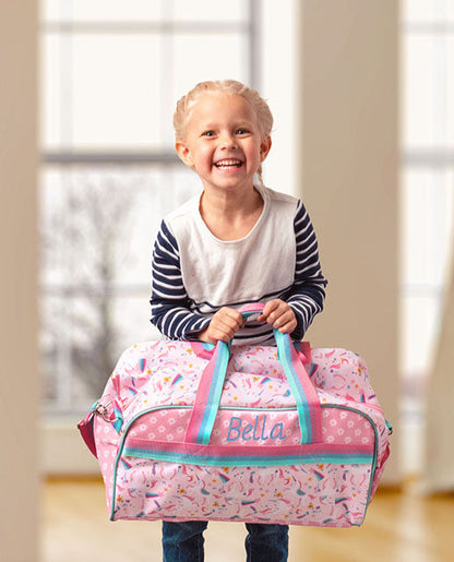 Pink unicorn-patterned duffle bag with turquoise handles and embroidered name 'Bella' held by a smiling child.