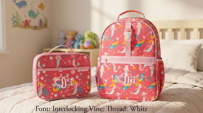 Pink mermaid all-over print Stephen Joseph backpack and lunch bag with white embroidered "M.D.L." monogram, set on a bed