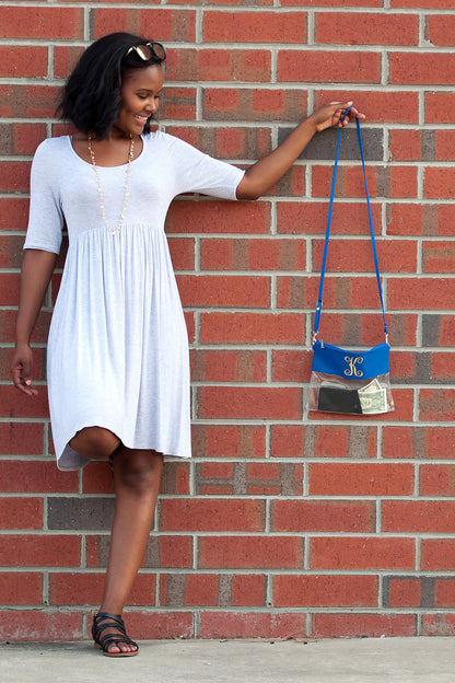 Clear bag with royal blue trim, held by its long strap beside a brick wall