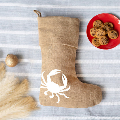Natural burlap Christmas stocking with a white crab design beside a plate of cookies