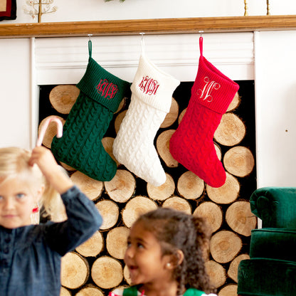 Green, cream and red monogrammed cable knit stockings above stacked firewood with children nearby