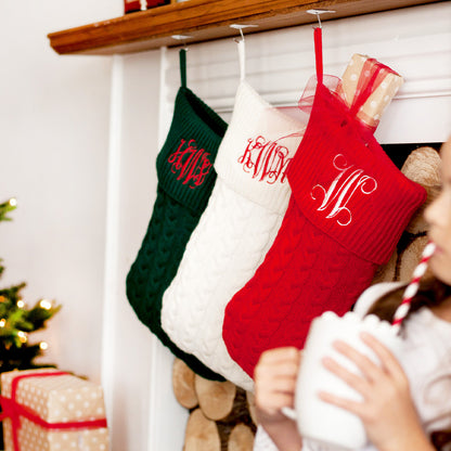 Green, cream and red cable knit Christmas stockings with embroidered monograms hanging from a mantel