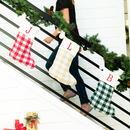 Red, tan and green check knit Christmas stockings with cream cuffs and embroidered initials hanging from a stair railing