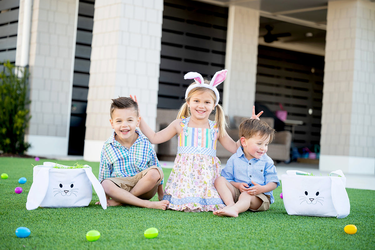 Two white plush bunny baskets on a lawn with three children and scattered Easter eggs