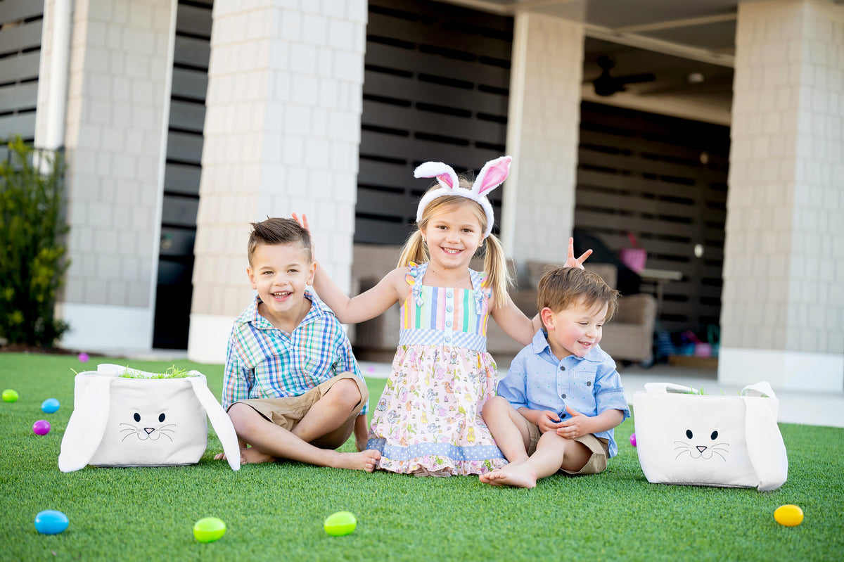 Two white plush bunny baskets with embroidered faces flanking three children on a lawn with colorful Easter eggs