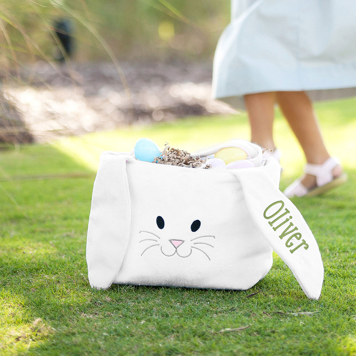 White plush bunny basket filled with pastel eggs on grass, personalized with the embroidered name Oliver on its ear