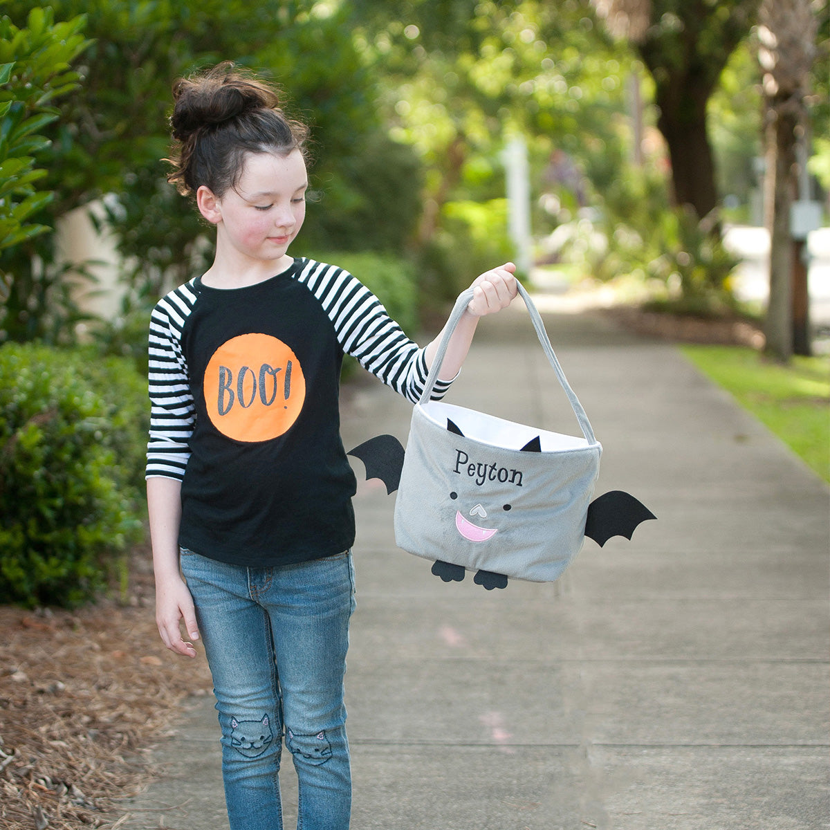 Girl in a BOO shirt holding a gray plush bat basket with black wings, ears, and the embroidered name Peyton
