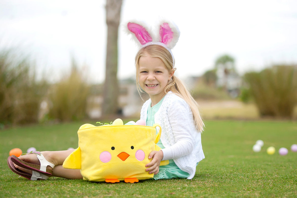 Girl in bunny ears holding a yellow plush chick basket filled with Easter eggs on grass