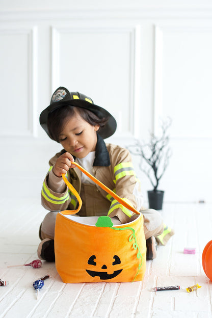 Child in a firefighter costume sitting on a white floor holding a plush orange pumpkin basket with scattered candy