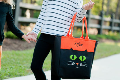 Woman walking with a black cat character tote with green eyes, orange panel embroidered Kylie in black, and orange handles