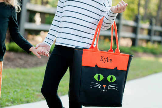 Woman walking with a black cat character tote with green eyes, orange panel embroidered Kylie in black, and orange handles