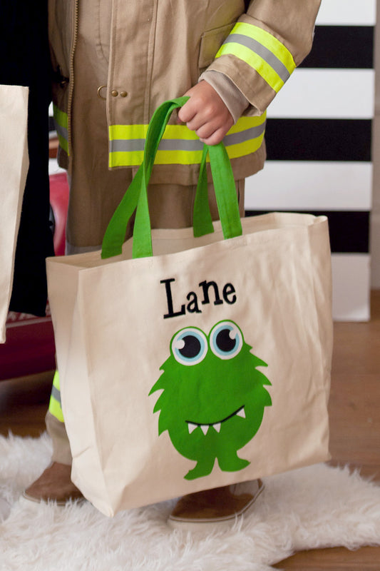 Child in a firefighter costume holding a cream canvas tote with a green monster, blue eyes, and the name Lane