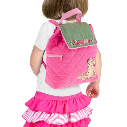 Toddler wears quilted pink Stephen Joseph backpack with green floral flap, leopard appliqué, and pink straps over white shirt and ruffled skirt