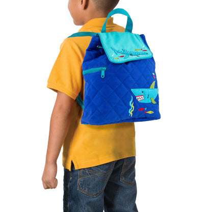 Toddler wearing blue quilted Stephen Joseph backpack with embroidered shark print, teal flap, and yellow shirt with blue jeans