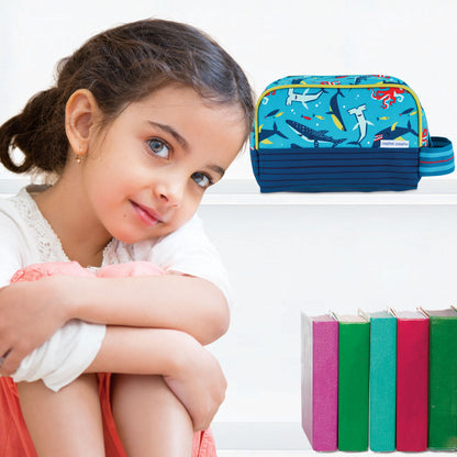 Young girl resting chin on crossed knees beside white shelf holding blue Stephen Joseph Shark toiletry bag with shark print and striped handle
