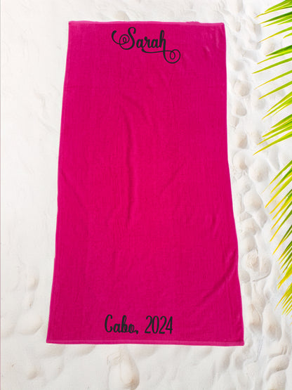 Hot pink cotton velour beach towel on white sand with black script 'Sarah' and 'Cabo, 2024' text.