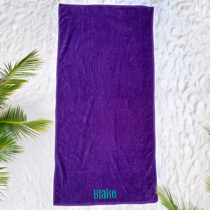 Purple solid color beach towel with the name Blake embroidered in teal at the bottom, laid on white sand.