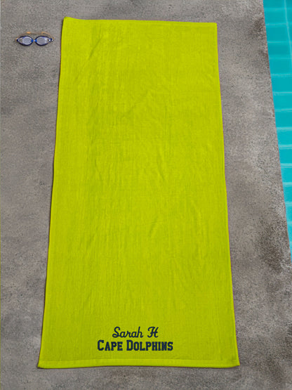 Lime green beach towel with embroidered text 'Sarah H CAPE DOLPHINS' lying on concrete near a pool.