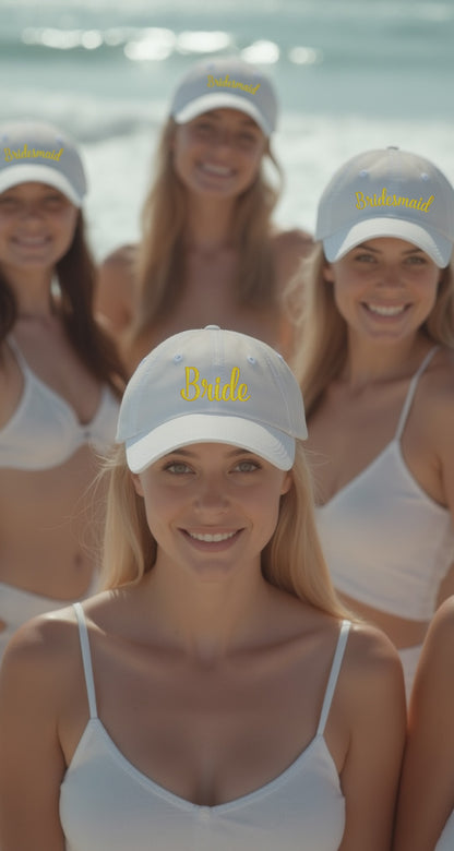 White unstructured dad hats with yellow embroidered 'Bride' and 'Bridesmaid' text, worn by women on a beach.