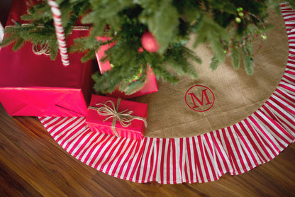 Natural burlap Christmas tree skirt with red and white striped ruffle and embroidered monogram beneath a tree