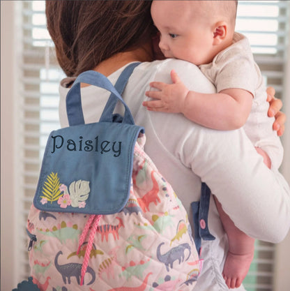 A baby rests on a person's shoulder beside a personalized Stephen Joseph pink dinosaur quilted backpack with blue pocket embroidered "Paisley"