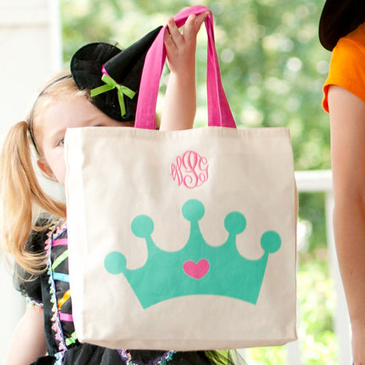 Girl in a witch costume holding a cream canvas tote with teal crown, pink heart, and pink script monogram