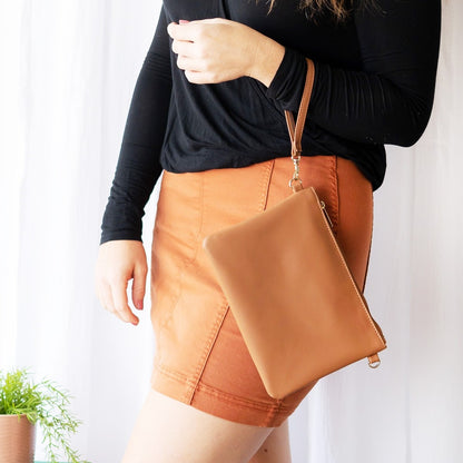 Tan leather wristlet with a matching strap, held by a person wearing a black top and rust-colored skirt.