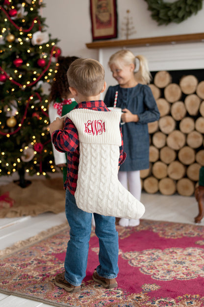 Child holding a cream cable knit stocking with red embroidered monogram beside a Christmas tree