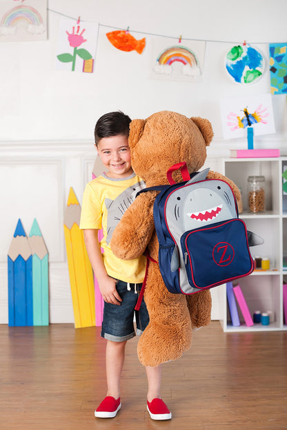 Child carrying a teddy bear beside a shark backpack indoors