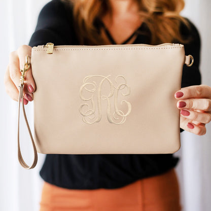 Beige leather wristlet with gold hardware and a large embroidered script monogram, held by a woman.