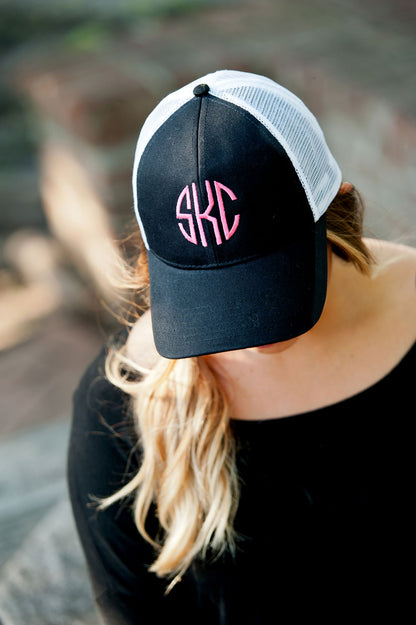 Woman wearing a black trucker hat with a pink monogram