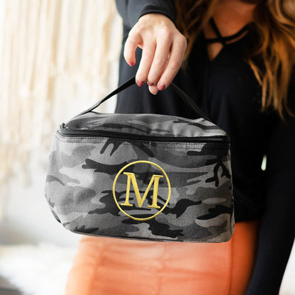 Gray camouflage cosmetic case with a yellow monogram held by its top handle