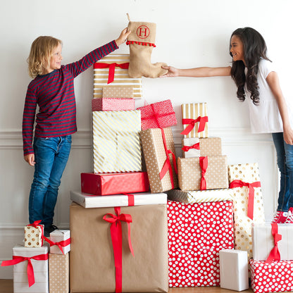 Two people holding a natural Christmas stocking with red pom-pom trim above stacked gifts