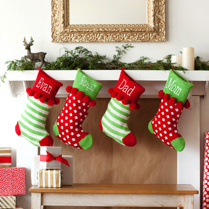 Green striped and red dotted knit stockings with embroidered names and pom-pom cuffs hanging from a mantel