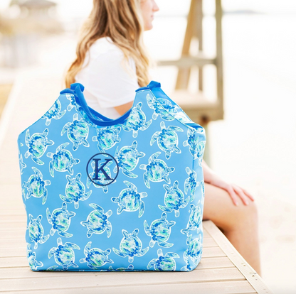Blue turtle-print beach bag with a dark monogram beside a seated woman