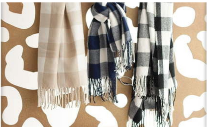 Three fringed plaid scarves in beige, navy, and black hang against a brown and white abstract patterned background.