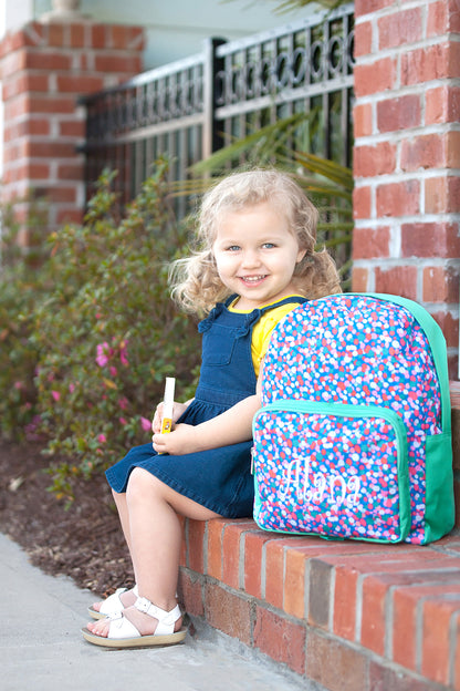 Child sitting beside a confetti-print preschool backpack on a brick wall