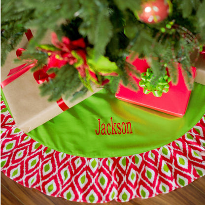 Green Christmas tree skirt with red and white ikat border and embroidered name beneath a decorated tree